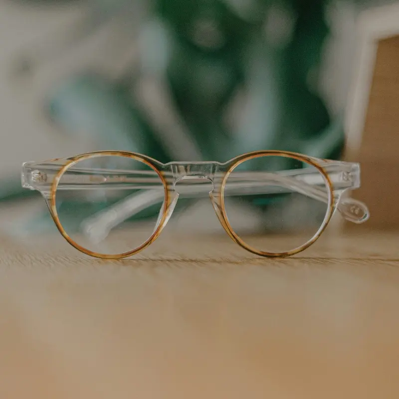 Clear frame round glasses with tortoiseshell lens rims and clear lenses resting on a wooden surface with blurred green plant in background