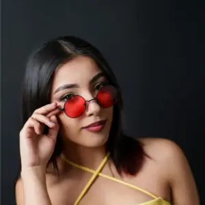 Woman holding small round metal sunglasses with red lenses in front of her face against a dark background