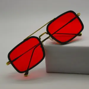 Black and gold double-bridge square sunglasses with vivid red lenses leaning against a white display block on a grey background