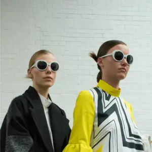 Two models wearing oversized white round injection-molded sunglasses with dark lenses against a white brick wall