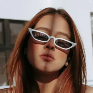 Woman wearing white narrow cat-eye sunglasses with brown lenses in bright sunlight against a light background