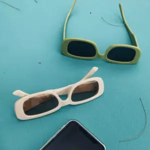Two injection-molded PC sunglasses on a teal background — olive green wide-frame pair with dark lenses and cream narrow-frame pair with gradient brown lenses