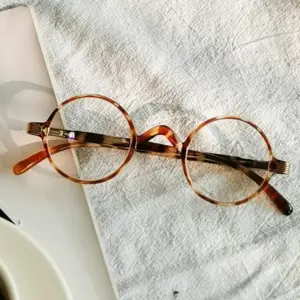 Round amber tortoiseshell acetate reading glasses on a textured fabric surface, representing a brand's decision on lens options like blue light