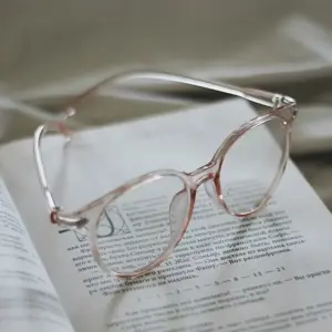 Clear pink translucent acetate reading glasses resting on an open book, illustrating frame material choice for reading glasses brands
