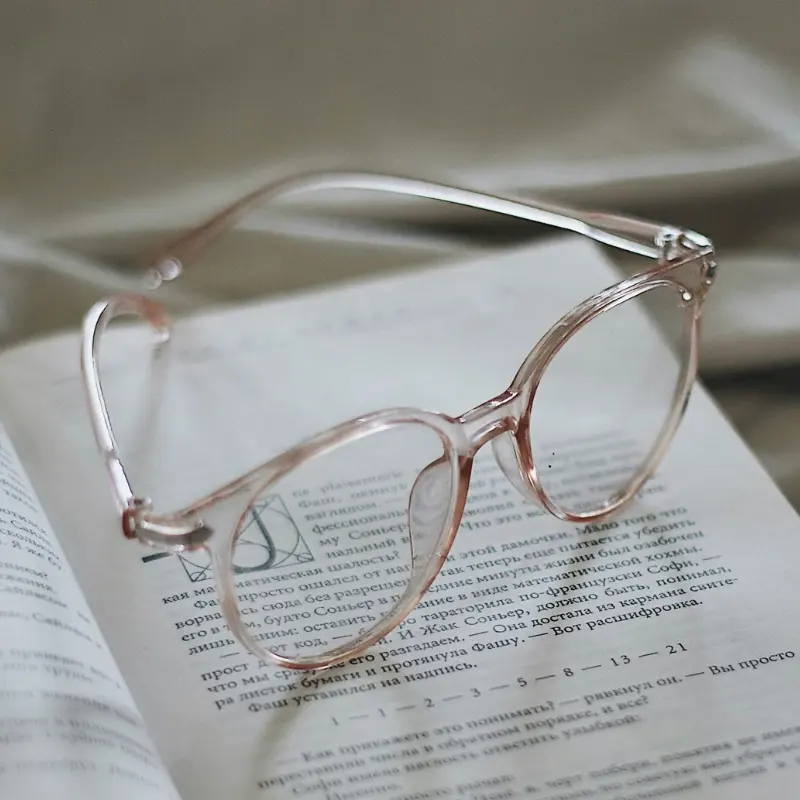 Clear pink translucent acetate reading glasses resting on an open book, illustrating frame material choice for reading glasses brands