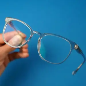 Hand holding transparent light blue square-round frame glasses with clear lenses showing a subtle blue-green surface reflection against a blue background