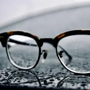 Close-up of tortoise acetate browline eyeglasses with thin metal lower rim, raindrops on the lens in a moody outdoor setting
