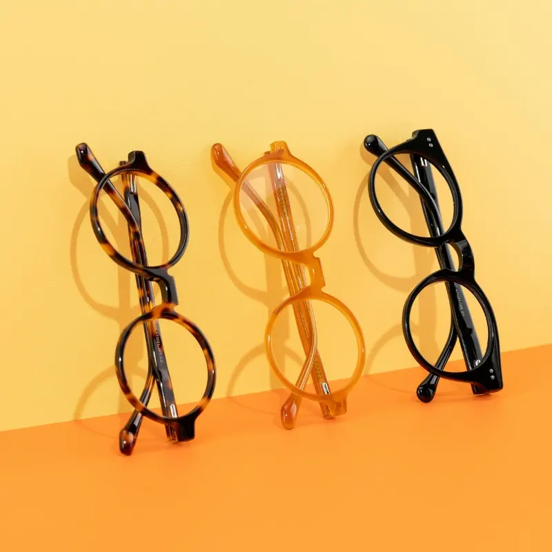 Three round acetate optical frames in tortoise, amber, and black standing upright against a yellow and orange background